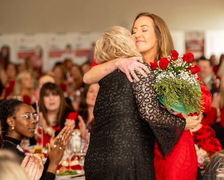 Two women hugging while one holds a bouquet of red roses.